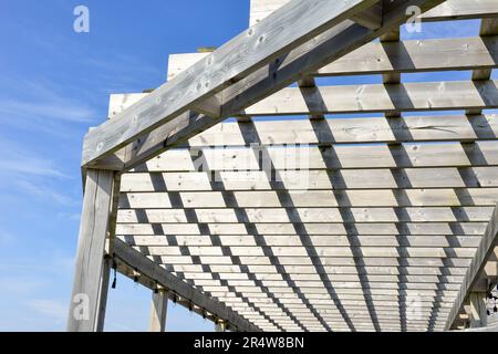 A grey colored worn and weathered wood pergola roof with a blue sky in the background. The outdoor sun shelter has wooden slats hanging over the wood Stock Photo