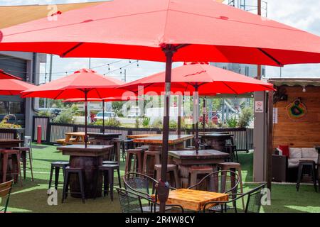 An empty brewery with multiple round shaped red nylon sunshades or patio umbrellas over a patio deck of black metal chairs and pub tables. Stock Photo