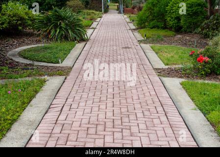 A red brick stone walkway through a lush green garden of lush green grass, shrubs, trees, and colorful flowers. There's concrete edging along the path. Stock Photo