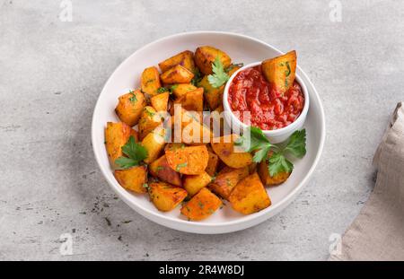 Traditional Spanish potato, patatas bravas with smoked paprika, spicy tomato sauce and parsley in a white bowl on a gray stone background, top view. D Stock Photo