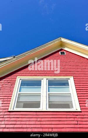 The outside of a peaked exterior wall of a house with vibrant red siding with white trim or edge. There's a small glass window with shades in the top Stock Photo