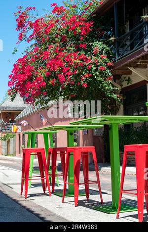 Vibrant red and colorful green colored metal pub-size tables and chairs are on a sidewalk in front of a coffee shop with trees and red flowers bloom Stock Photo