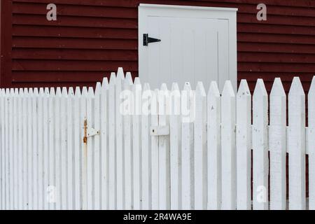 The exterior of a red wooden cape cod clapboard or batten board siding wall of a country style barn with a white wooden picket fence in the foreground Stock Photo