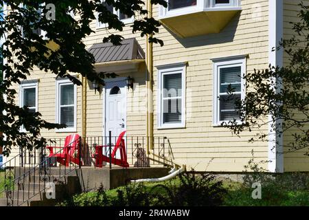 The exterior of a vintage yellow colored wooden clapboard country style house with red Adirondack chairs on the front patio. Stock Photo