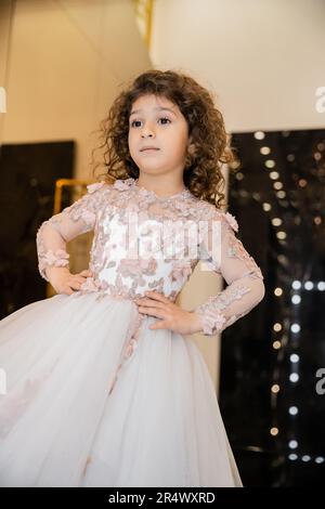 curly middle eastern and little girl in floral dress with tulle skirt standing with hands on hips and looking away in bridal boutique, preparation for Stock Photo