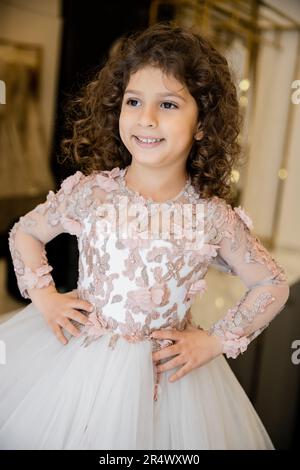 joyous middle eastern and little girl in floral dress with tulle skirt standing with hands on hips and looking away in bridal boutique, preparation fo Stock Photo