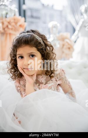 portrait of cute middle eastern little girl with curly hair sitting in floral dress on white couch inside of luxurious wedding salon, tulle skirt, blu Stock Photo