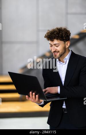 Young modern businessman wearing blue blazer and casual shirt, standing with open laptop in hands Stock Photo