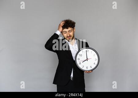 Shocked worried young bearded business man 20s in classic white shirt standing hold clock spreading hands isolated on grey color wall background studi Stock Photo