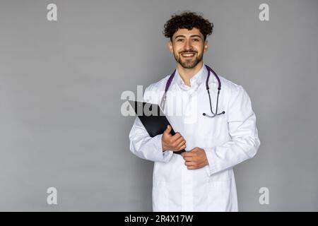 Happy caucasian doctor with stethoscope holding clipboard and writing something while standing and smiling isolated on grey background Stock Photo