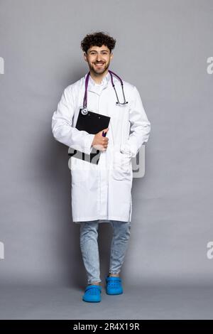 Happy caucasian doctor with stethoscope holding clipboard and writing something while standing and smiling isolated on grey background Stock Photo