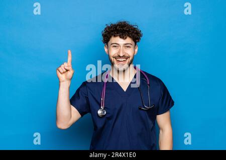 Young handsome doctor man with beard wearing coat and glasses over blue background pointing finger up with successful idea. Exited and happy. Number o Stock Photo