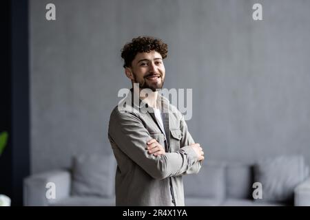 Happy confident millennial businessman in casual standing with arms crossed at work table in home office, looking at camera, smiling. Male freelance p Stock Photo