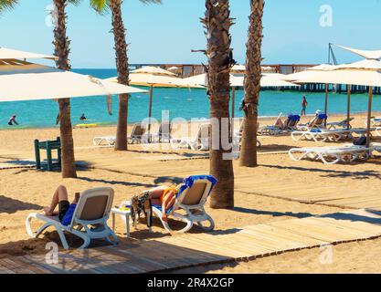 Antalya; Turkey-September 10; 2022: Tourist couple resting on sunbeds and sunbathing under the palm trees on the beach. People swimming in the sea on Stock Photo