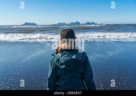 View taken from behind of a close-up of a woman standing on the south coast of Iceland looking out to sea at the Westman Islands Stock Photo