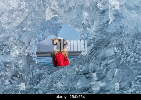 Close-up view through a round, clear ice formation of a woman wearing a woolen hat looking out and admiring the icebergs along the South Coast of I... Stock Photo