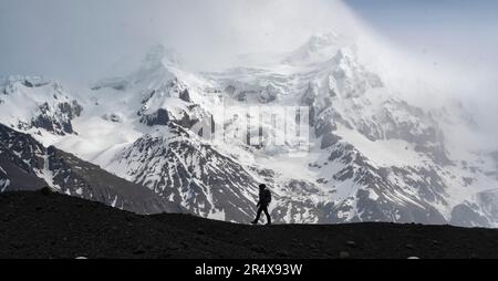 Silhouette of a person walking along a glacial moraine in front of a large snow-capped mountain on the South Coast of Iceland; South Iceland, Iceland Stock Photo