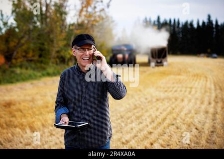 Farmer using a tablet to manage his harvest and talking on his cellphone with harvesting equipment working in the background Stock Photo