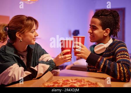 Side view portrait of retro young couple eating pizza and toasting with red paper cups Stock Photo