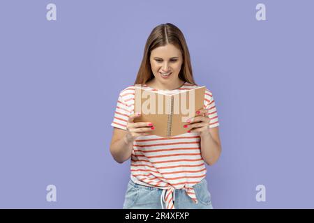 Portrait of satisfied smiling woman wearing striped T-shirt holding and reading book, being very interested in plot, expressing positive emotions. Indoor studio shot isolated on purple background. Stock Photo