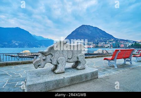 LUGANO, SWITZERLAND - MARCH 14, 2022: The stone statue of Rhinoceros in Park of Sculptures on embankment of Lake Lugano with Monte San Salvatore in ba Stock Photo