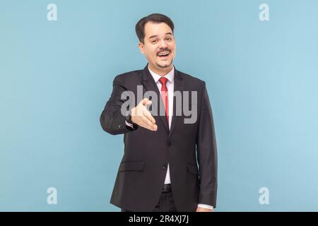 Portrait of friendly positive optimistic man with mustache standing outstretching hand, nice to meet you, wearing black suit with red tie. Indoor studio shot isolated on light blue background. Stock Photo