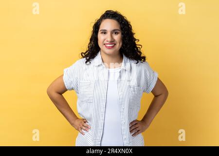 Portrait of friendly positive woman with dark wavy hair standing with hands on hips, looking at camera with satisfied facial expression. Indoor studio shot isolated on yellow background. Stock Photo