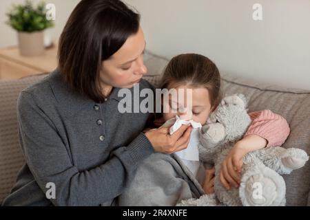 Sad worried caucasian mature mother blows nose to napkin to little daughter, lies on sofa in living room Stock Photo