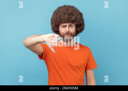Portrait of displeased man with Afro hairstyle wearing orange T-shirt showing thumbs down dislike gesture, symbol of disagree, giving feedback. Indoor studio shot isolated on blue background. Stock Photo