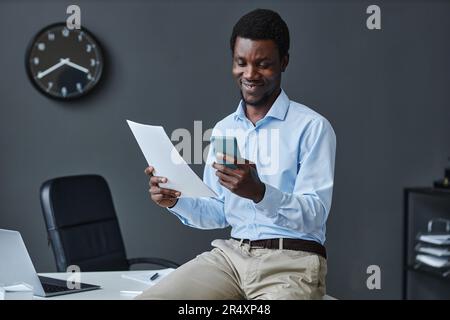 Portrait of young black businessman using smartphone sitting on desk in office and smiling, copy space Stock Photo