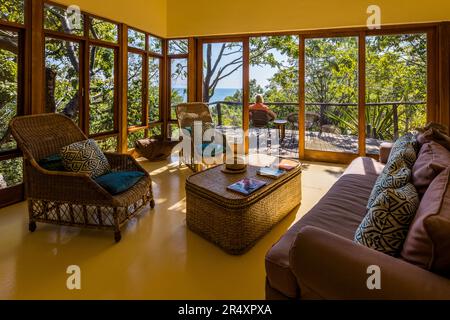 The living room of a detached villa at Pumulani Lodge (Robin Pope Safaris) looks through floor-to-ceiling windows onto Lake Malawi near Cape Maclear, Lake Malawi National Park, Malawi; on the terrace sits Angela Berg. Stock Photo