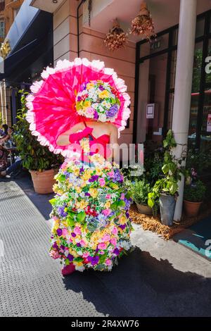A woman wearing an elaborate colourful floral dress carrying a red parasol by the Sloane Square Hotel, Chelsea, London SW1 during Chelsea in Bloom Stock Photo