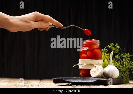 Pickled cherry tomatoes in an open transparent jar on a dark wooden background, one canned tomato on a vintage fork in a female hand. Stock Photo