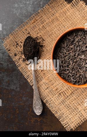 Dry black tea leaves in a clay bowl and a vintage spoon on a dark wooden background top view. Large-leaf and small-leaf black tea. Stock Photo