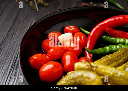 Canned cherry tomatoes, cucumbers, chili peppers in a round black plate on a dark background, close-up. Mix of pickled vegetables, spices Stock Photo