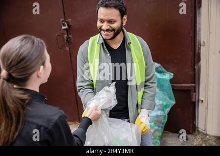 Smiling indian worker in safety vest and gloves taking trash bag from blurred volunteer in outdoor waste disposal station, garbage sorting and recycli Stock Photo