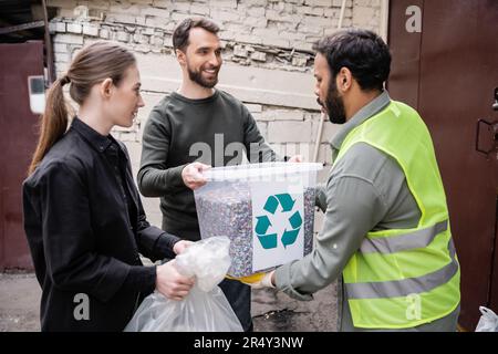 Smiling volunteers giving trash to indian worker in high visibility vest in outdoor waste disposal station, garbage sorting and recycling concept Stock Photo