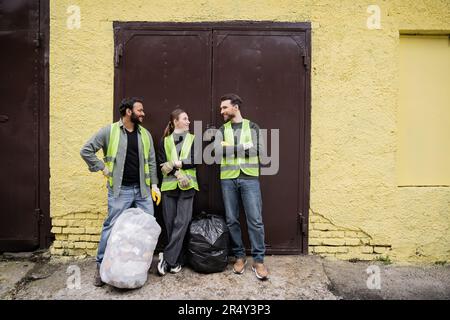 Smiling multiethnic workers in protective vests and gloves talking near trash bags and door of waste disposal station outdoors, garbage sorting and re Stock Photo