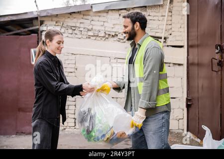 Positive indian worker in safety vest and gloves taking plastic bag with trash from volunteer outdoors in waste disposal station, garbage sorting and Stock Photo