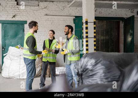 Smiling interracial workers in protective vests and gloves talking while standing and resting near blurred plastic bags with trash in garbage sorting Stock Photo