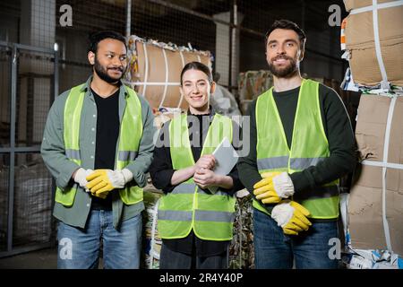 Smiling worker in protective vest holding digital tablet and looking at camera while standing near multiethnic colleagues and waste paper waste in dis Stock Photo