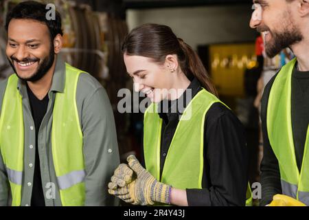Smiling young female worker in protective gloves and vest standing next to multiethnic colleagues while working together in blurred waste disposal sta Stock Photo