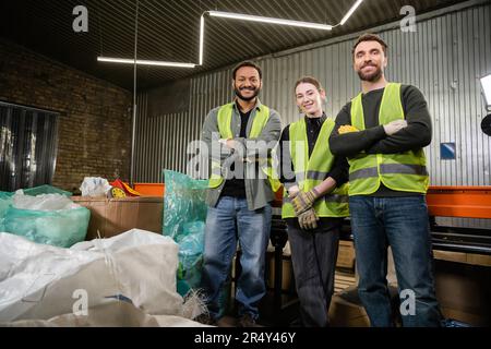 Cheerful multiethnic workers in safety vests and protective gloves looking at camera while standing near plastic bags and conveyor in waste disposal s Stock Photo