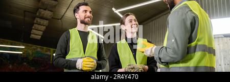Cheerful workers in protective gloves and high visibility vests looking at indian colleague talking while standing together in blurred waste disposal Stock Photo