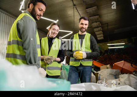 Smiling multiethnic workers in protective vests and gloves looking at blurred sacks with trash for recycle while working together in waste disposal st Stock Photo