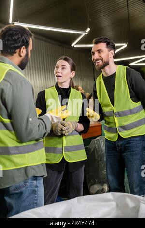 Positive workers in high visibility vests and gloves talking to indian colleague while standing near sack and working together in waste disposal stati Stock Photo