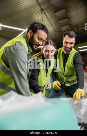 Smiling multiethnic workers in protective clothes and gloves talking and working near blurred sacks together in waste disposal station, garbage sortin Stock Photo