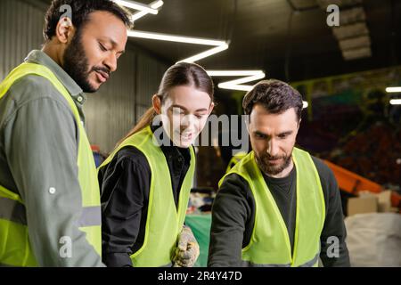 Cheerful workers in high visibility vests looking away while indian colleague talking and standing together in blurred garbage sorting center, garbage Stock Photo