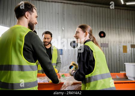 Smiling woman in protective uniform and gloves holding garbage and talking to interracial colleagues while working together near conveyor in waste dis Stock Photo