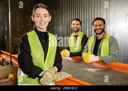 Smiling sorter in protective clothes and gloves looking at camera while standing near conveyor and blurred multiethnic colleagues in garbage sorting c Stock Photo